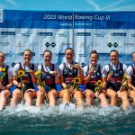 8 women and a male cox with gold medals sitting on landing stage with feet in water