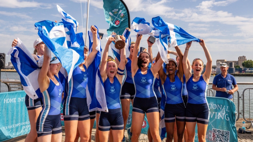 A group of rowers in blue celebrating on land with a trophy and flag